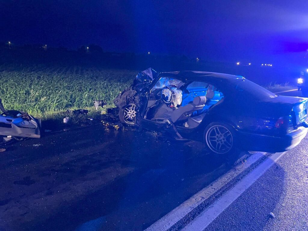 Uszkodzony samochód osobowy stoi na poboczu drogi, obok widać zniszczone elementy pojazdu, w tle migają niebieskie światła.
