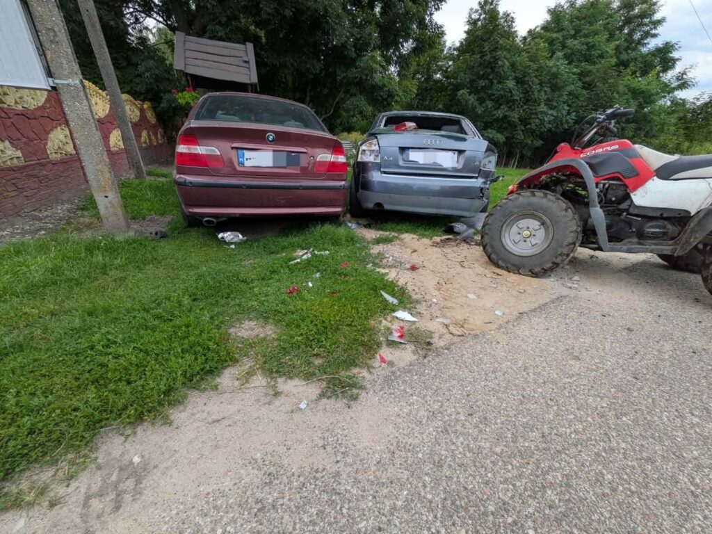 Dwa uszkodzone samochody zaparkowane obok siebie na trawie, obok quad z dużymi oponami. W tle widoczne drzewa.