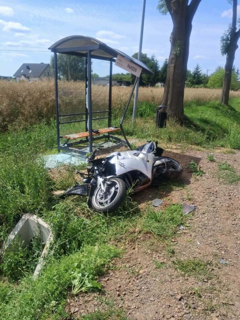 Zniszczony motocykl leży na trawie obok przystanku autobusowego z tabliczką "PARZEŃ" w otoczeniu roślinności.