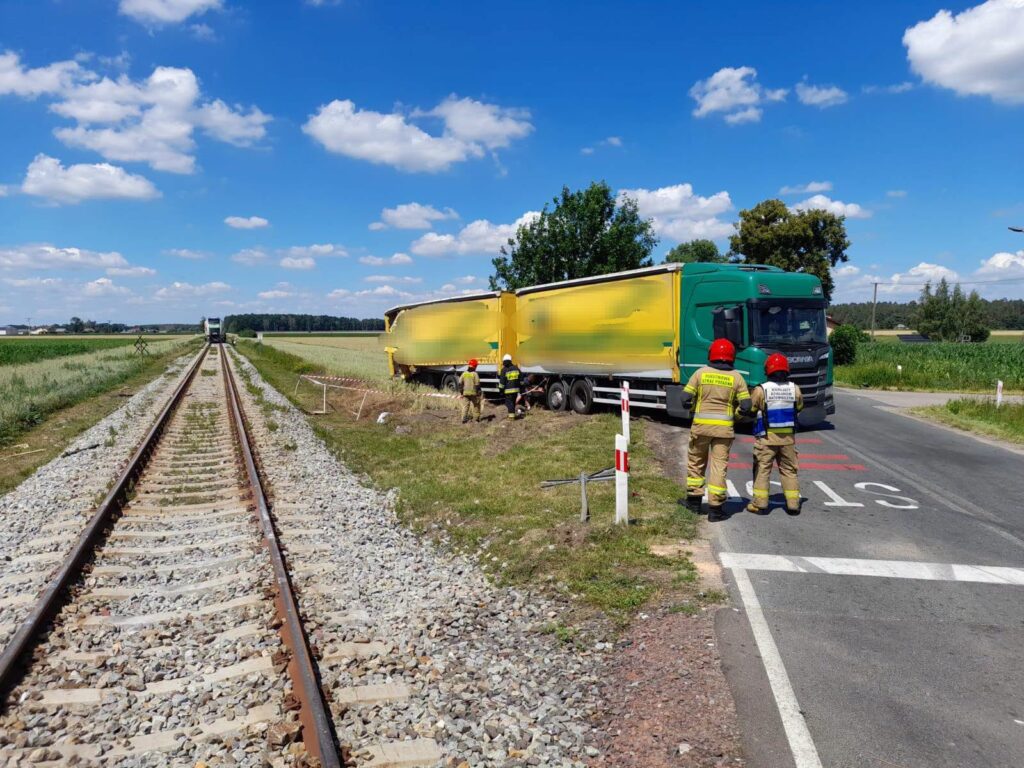 Strażacy w kaskach badają miejsce zderzenia ciężarówki z torami kolejowymi obok drogi, w tle widoczne pola.