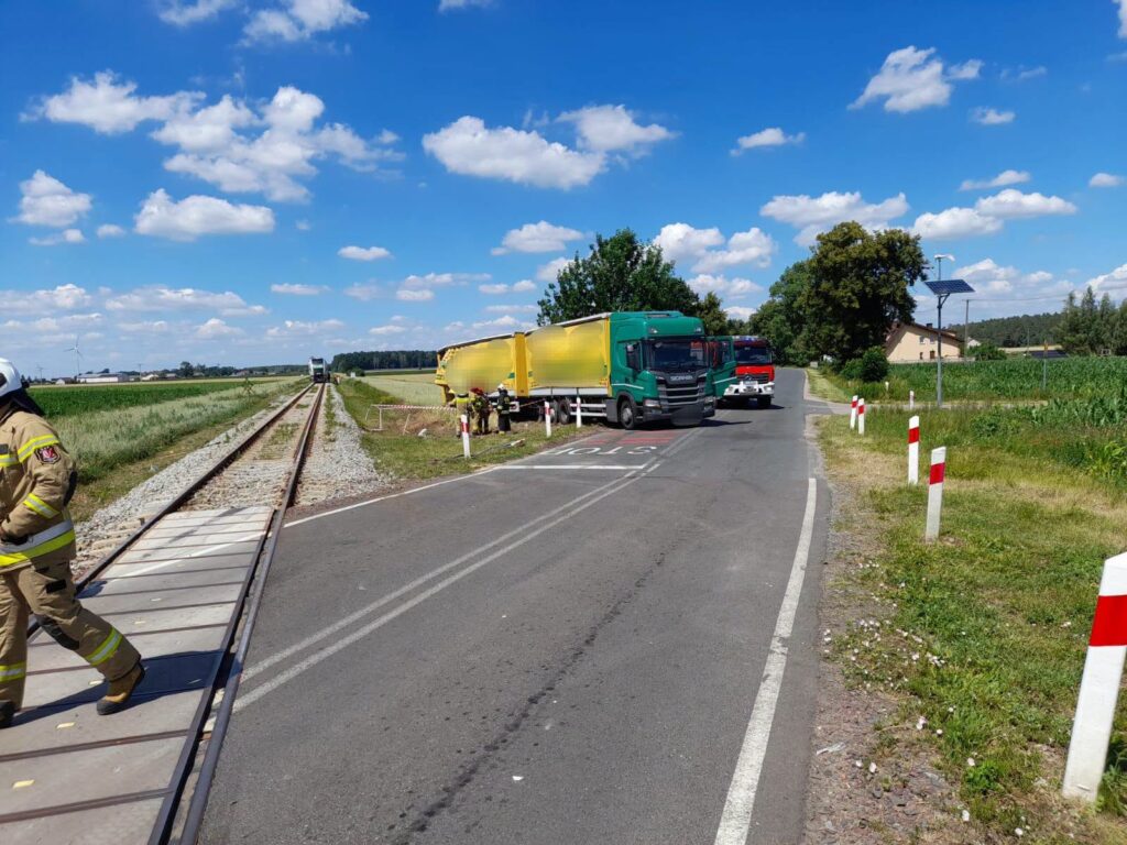 Strażak w mundurze idzie wzdłuż torów kolejowych, obok ciężarówki z żółtą naczepą zaparkowanej na drodze.