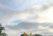 Czarny samochód osobowy z uszkodzoną karoserią stoi na poboczu drogi, a helikopter ratunkowy przelatuje nad okolicą.