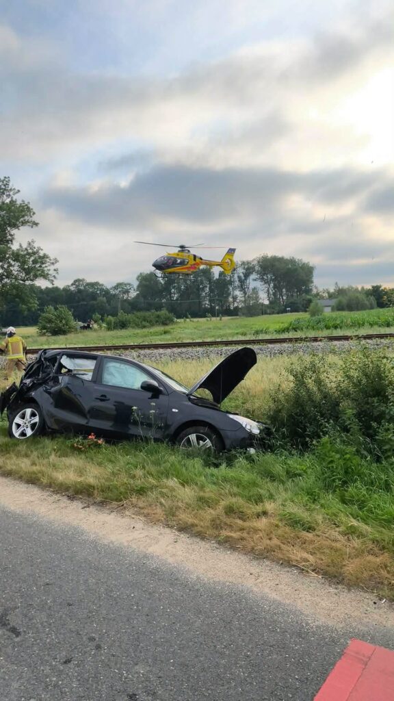Czarny samochód osobowy z uszkodzoną karoserią stoi na poboczu drogi, a helikopter ratunkowy przelatuje nad okolicą.