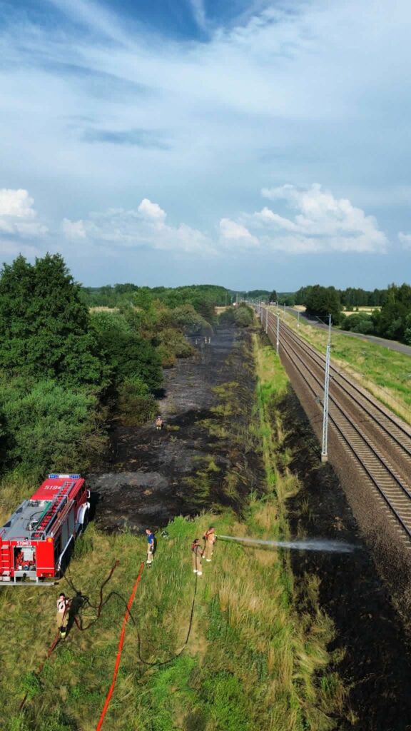 Strażacy gaszą pożar trawy obok torów kolejowych, używając węży strażackich i sprzętu gaśniczego.