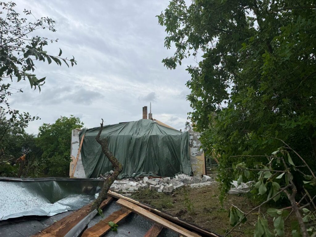 Uszkodzony budynek przykryty zielonym plandeką, z widocznymi zniszczeniami na ścianach i gruzem w otoczeniu.