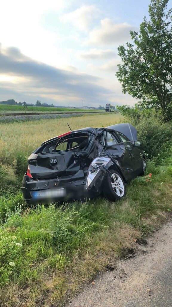Czarny samochód osobowy z uszkodzeniami leży w trawie obok drogi, w tle widoczne pola i tor kolejowy.