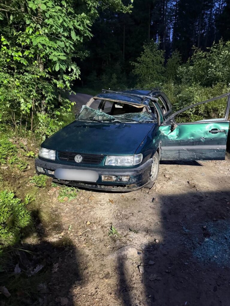 Zniszczony Volkswagen stoi na leśnej drodze, z otwartymi drzwiami i uszkodzoną karoserią, obok rosną drzewa.