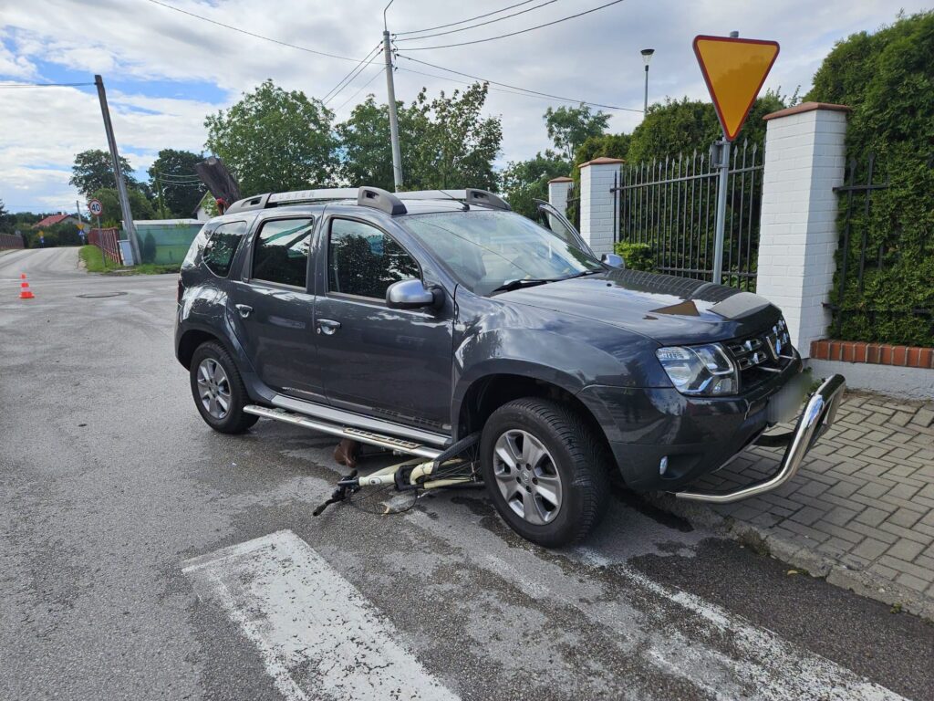 Czarny SUV zaparkowany na drodze z uszkodzeniami, obok leży rower, a w tle widoczne znaki drogowe i zieleń.