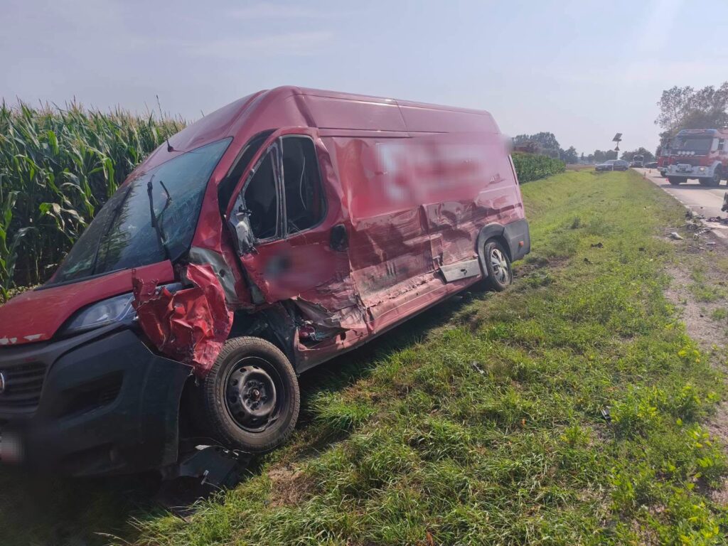 Czerwony furgon z uszkodzoną boczną częścią stoi na poboczu drogi, w tle widać pole kukurydzy i inne pojazdy.