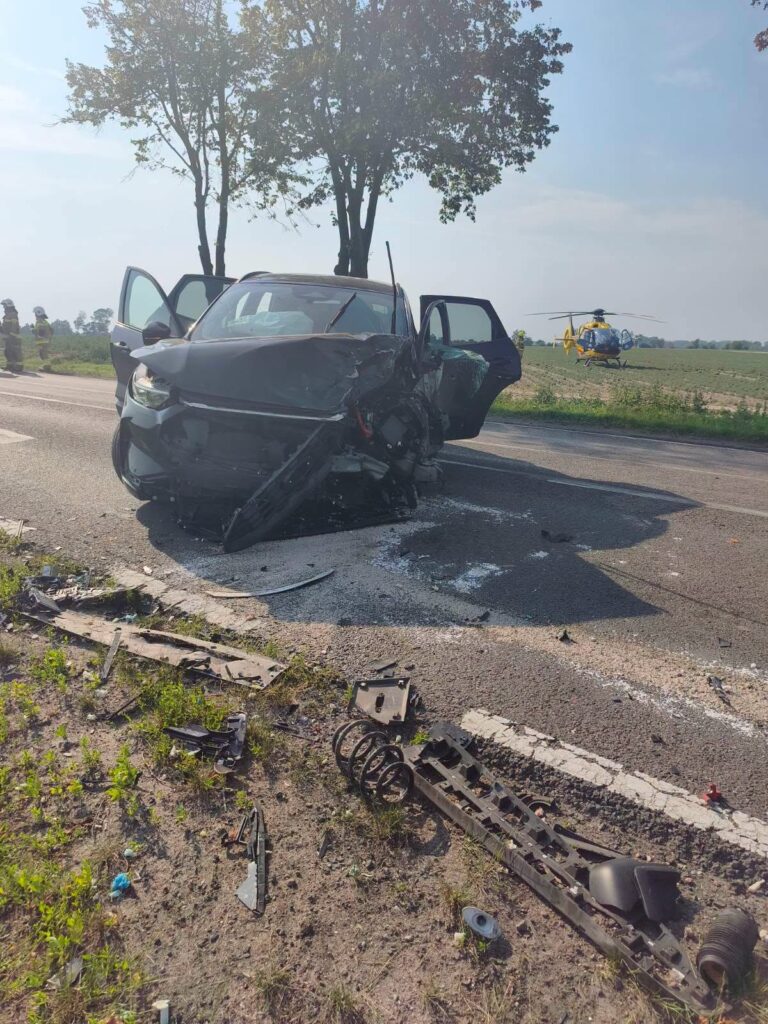 Rozbity samochód osobowy z uszkodzoną przednią częścią na drodze, obok leżące fragmenty pojazdów, w tle lądujący śmigłowiec LPR.