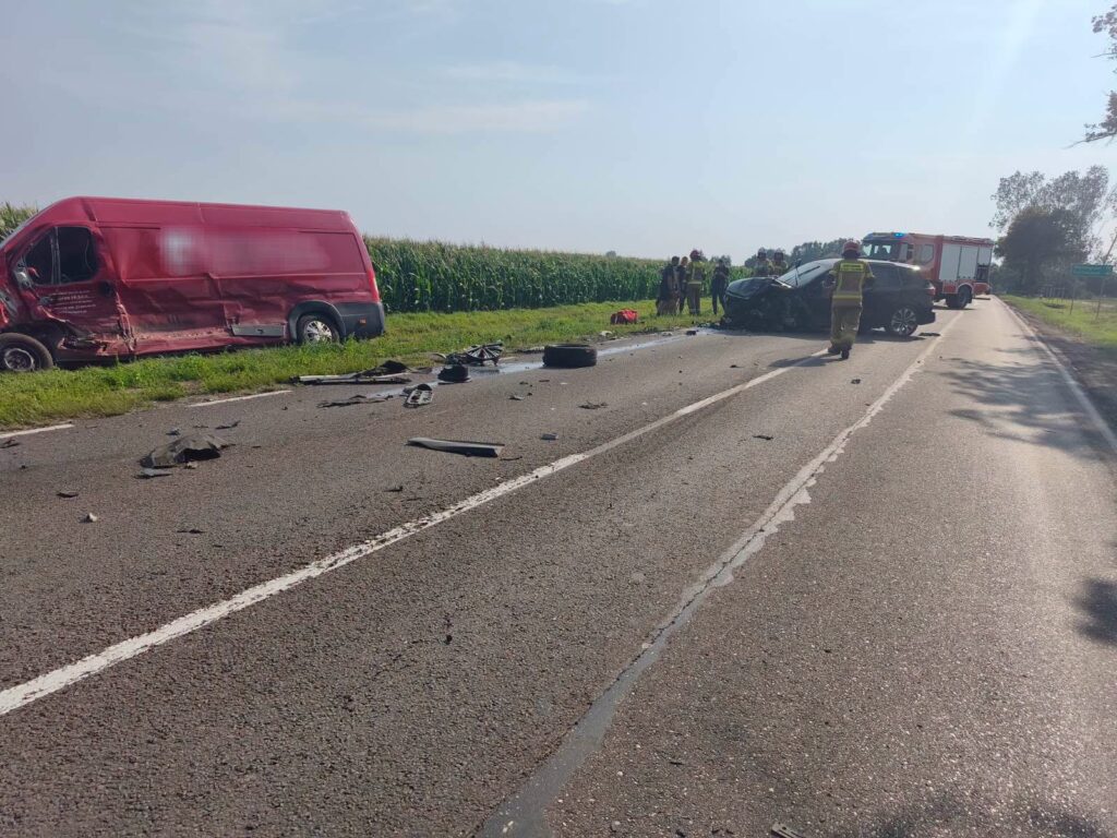 Wielki wypadek drogowy z udziałem czerwonawogo furgonu i osobowego samochodu na wiejskiej drodze, wokół pola kukurydzy.