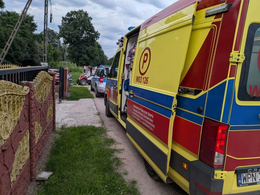 Zespół ratunkowy na miejscu zdarzenia, obok parkujących samochodów, w tle widoczni przechodnie.