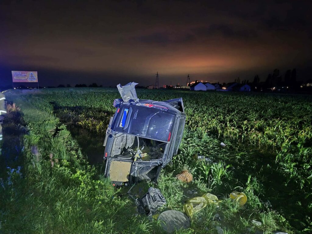 Samochód terenowy przewrócony na bok w polu, obok widoczne zboże oraz billboard w tle, nocne oświetlenie.