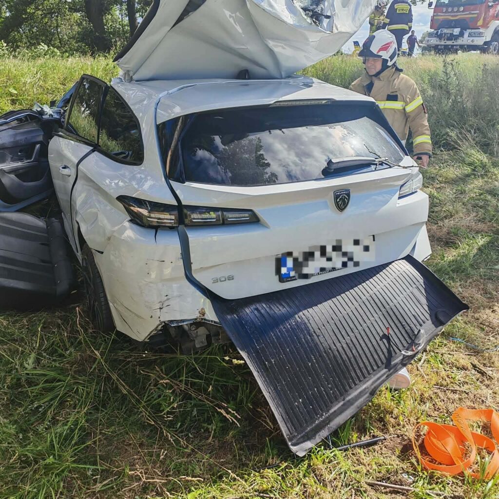 Rozbity samochód osobowy z odgiętą pokrywą bagażnika na tle zielonej trawy, obok strażak w hełmie.