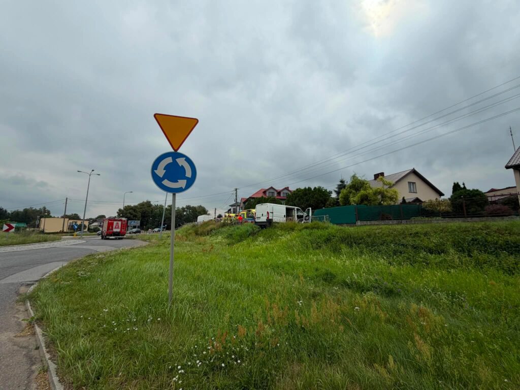 Rondo z oznaczeniem drogi okrężnej, w tle pojazdy i domy, nadchodzi pochmurna pogoda.