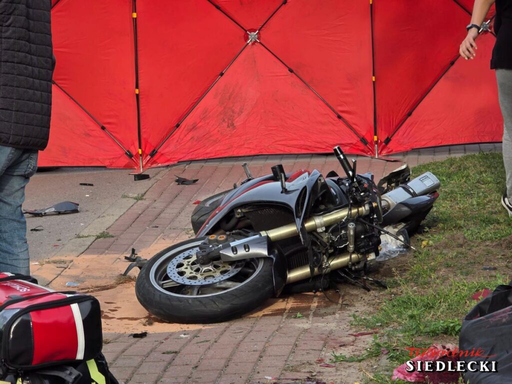 Motocykl leży na drodze, obok widoczne są uszkodzone elementy pojazdu oraz czerwony namiot zabezpieczający miejsce zdarzenia.
