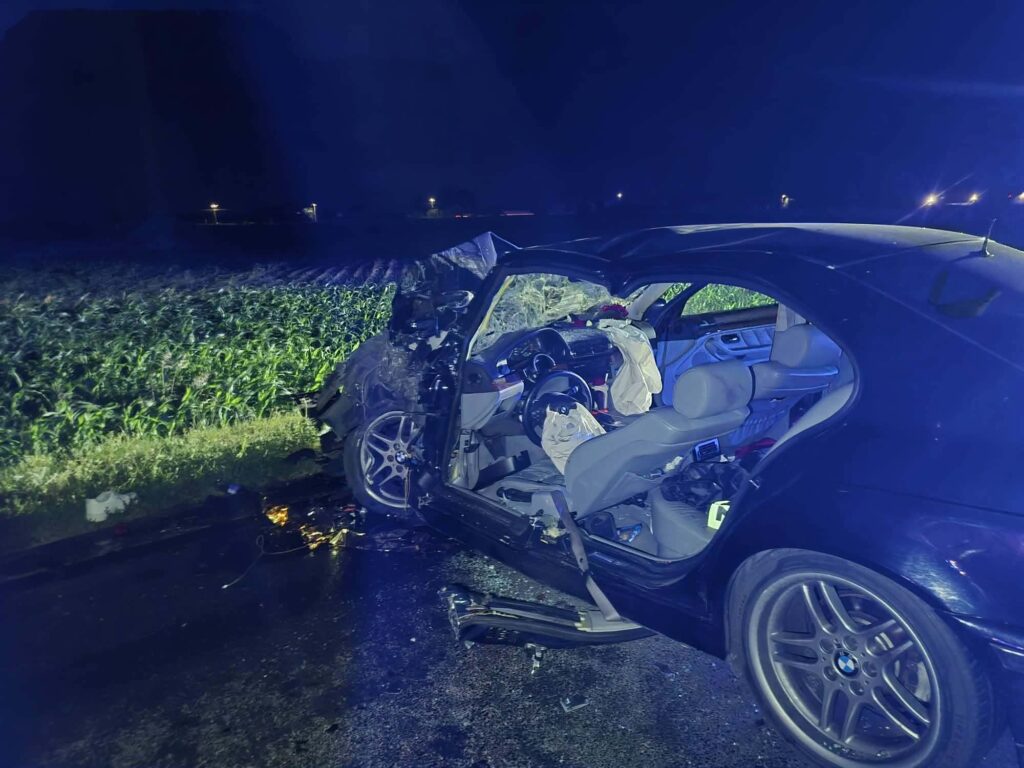 Zniszczony samochód osobowy leży na poboczu drogi, otoczony przez pole, z otwartymi drzwiami i wystrzelonymi poduszkami powietrznymi.