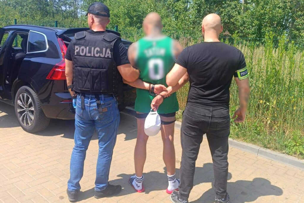 Mężczyzna w zielonej koszulce jest zatrzymywany przez dwóch policjantów w mundurach, obok czarnego SUV-a.
