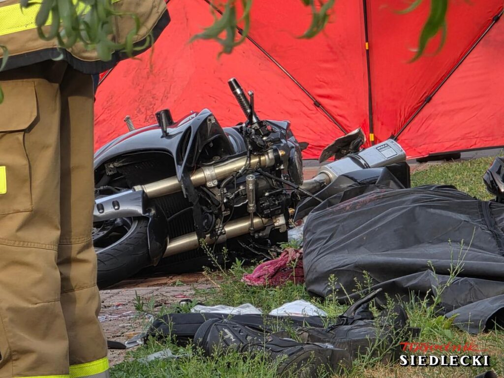 Motocykl leży na ziemi obok czarnego płótna, a w tle widoczny jest czerwony namiot ratunkowy.
