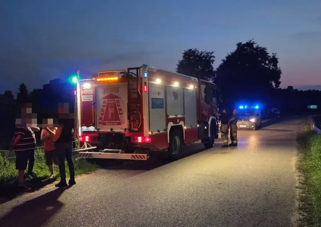 Grupa osób rozmawia obok wozu strażackiego na drodze, w tle widoczne niebieskie światła radiowozu policyjnego.