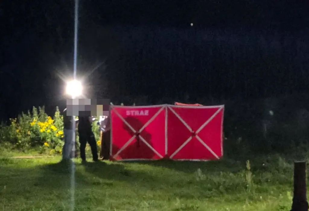 Grupa strażaków w mundurach rozmawia przy czerwonym namiocie na otwartej przestrzeni w nocy, oświetlonej przez reflektor.
