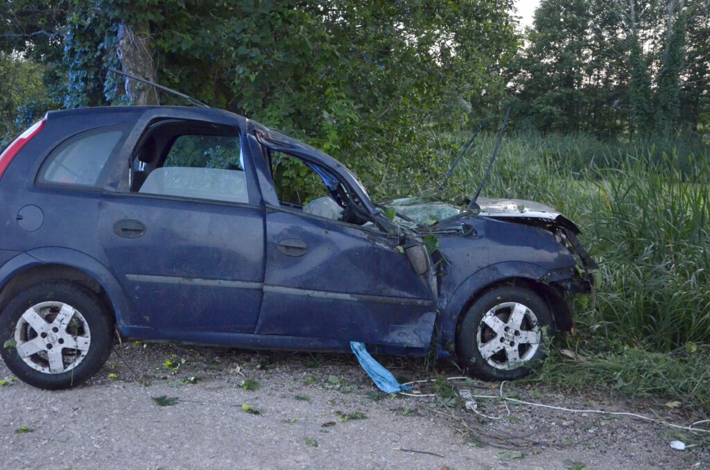 Uszkodzony niebieski samochód osobowy stoi na poboczu drogi, otoczony przez zarośla i drzewa.