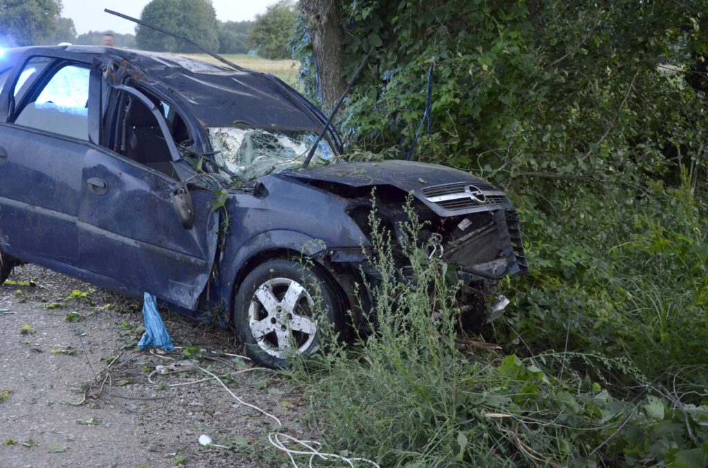 Uszkodzony niebieski samochód osobowy stoi w zaroślach przy drodze, z wgniecioną przednią częścią i rozbitą szybą.