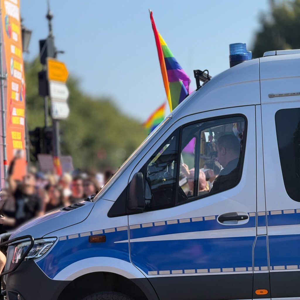 Funkcjonariusz policji w radiowozie z niebieską syreną, w tle tłum uczestników parady z flagami tęczowymi.