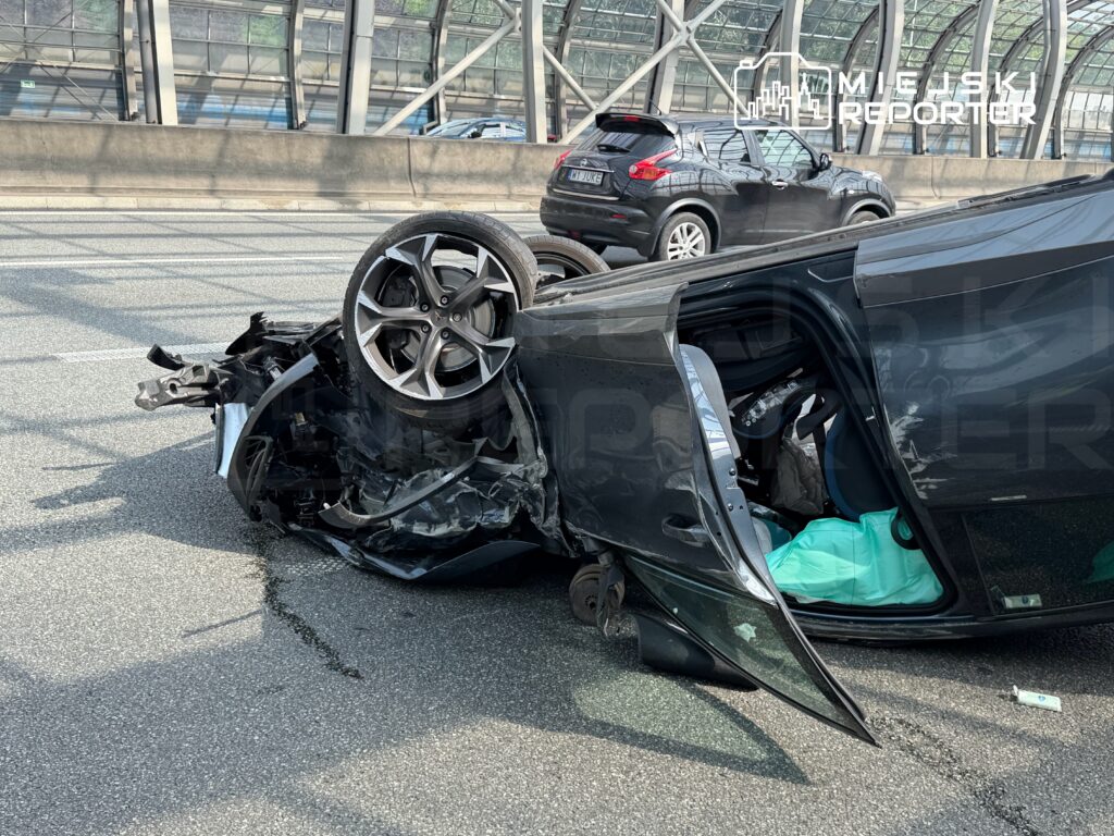 Zniszczony samochód leży na boku na pasie ruchu, z odwróconym kołem i otwartymi drzwiami.