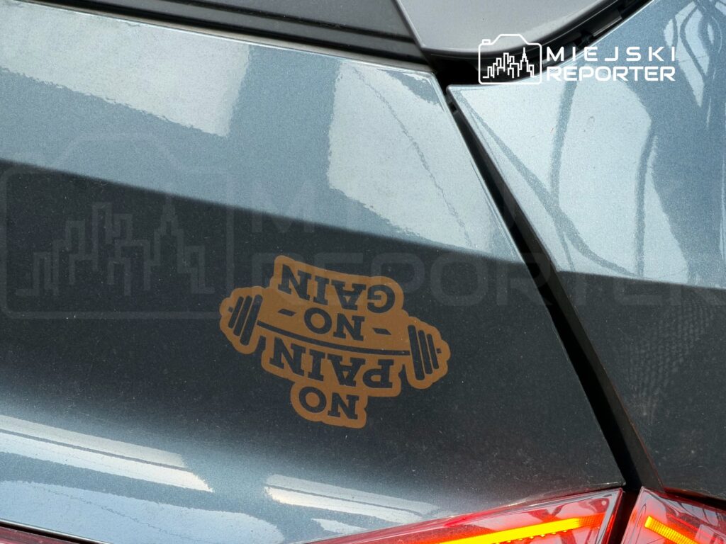 Na tylnej szybie samochodu znajduje się naklejka z napisem "NO PAIN NO GAIN" oraz grafiką ciężarka.