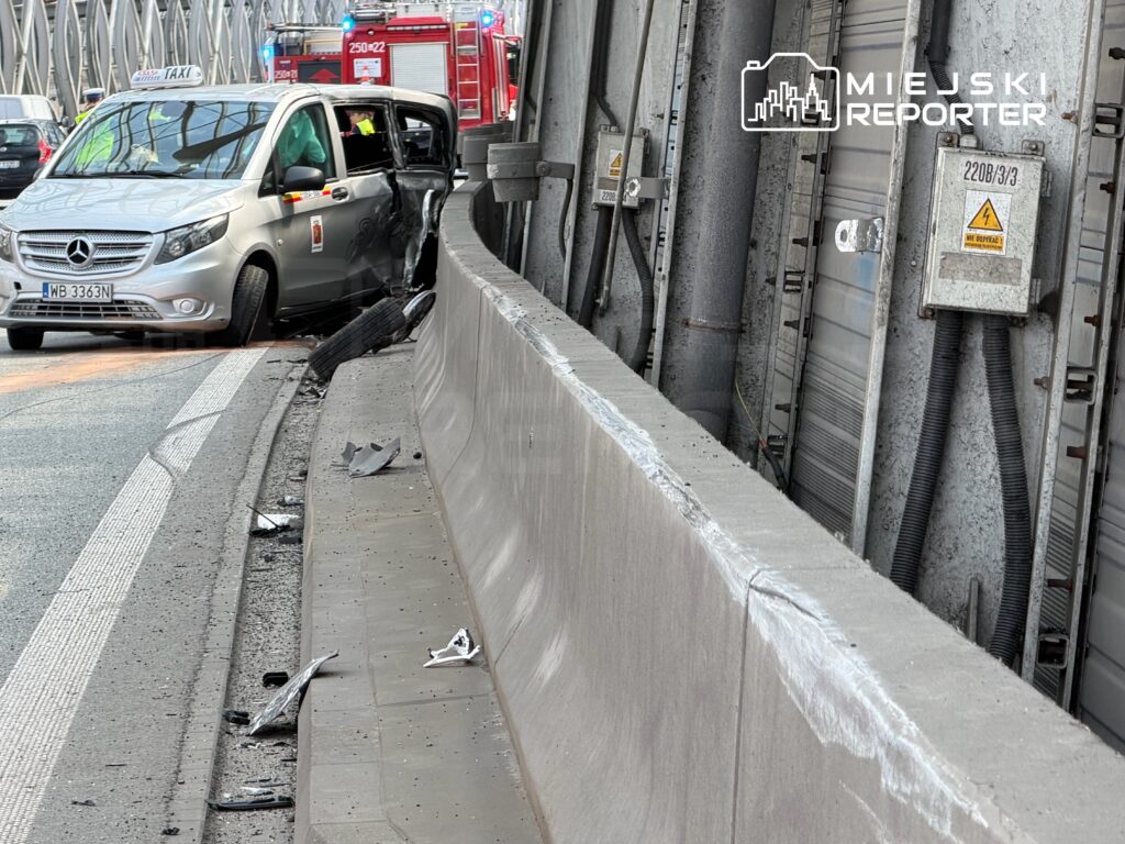 Wypadek samochodowy na drodze, gdzie uszkodzony taksówka stoi przy betonowym barierce, a obok leżą fragmenty wraku.