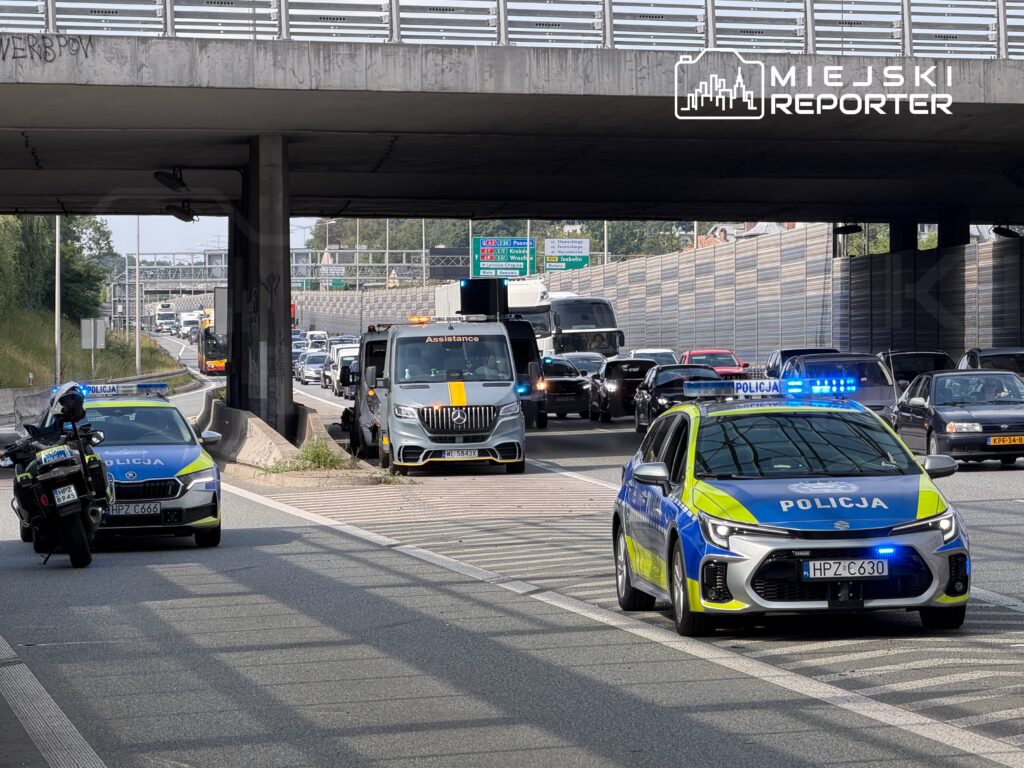 Policjanci w radiowozach kontrolują ruch na zatłoczonej drodze pod wiaduktem, obok stoi samochód pomocy drogowej.