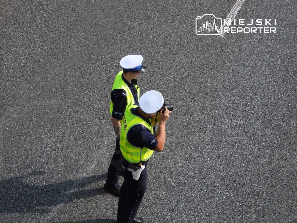 Dwaj policjanci w odblaskowych kamizelkach obserwują sytuację na drodze, jeden z nich robi zdjęcia.