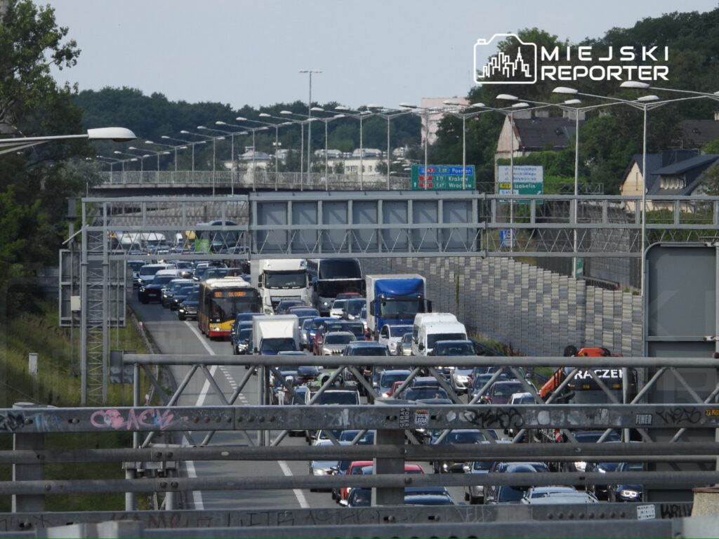 Ruch pojazdów w korku na wielopasmowej drodze, z widocznym autobusem i ciężarówkami, otoczony zielenią.