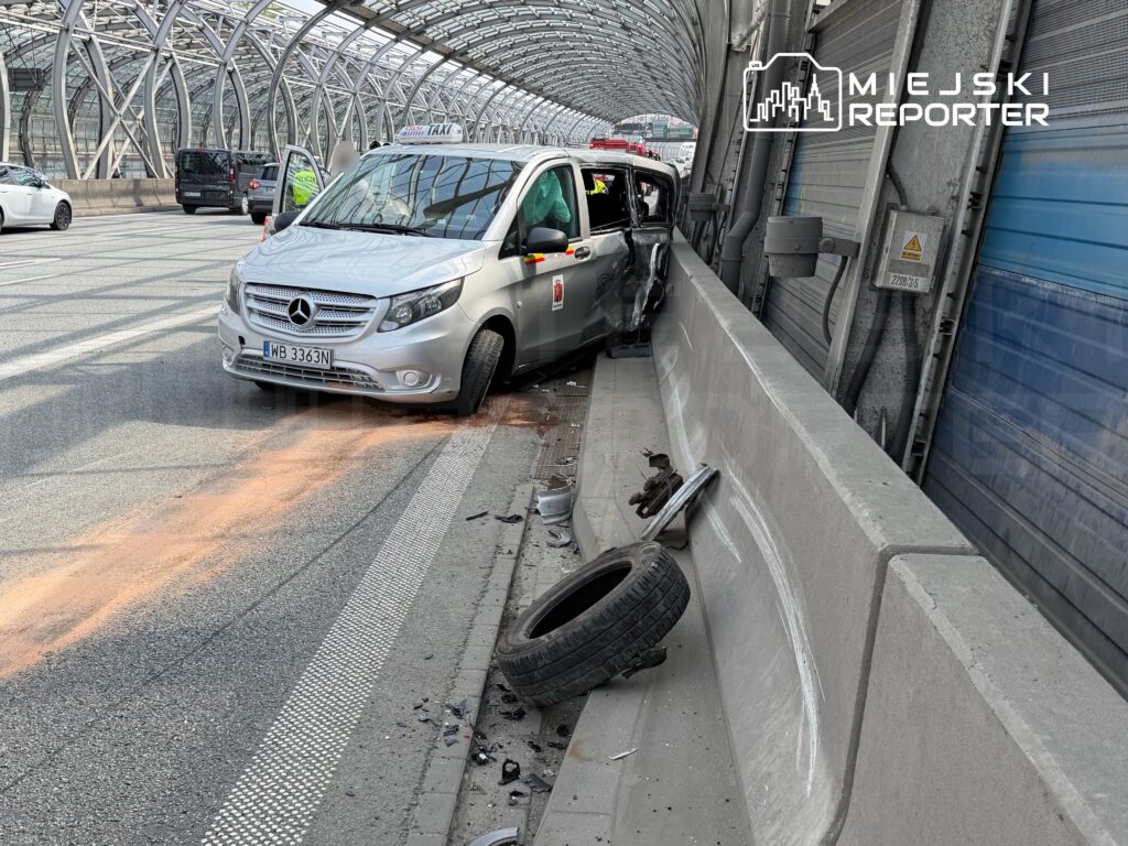 Taksi uderzone w barierkę ochronną na pasie ruchu, z uszkodzoną przednią częścią i odłamkami na jezdni.