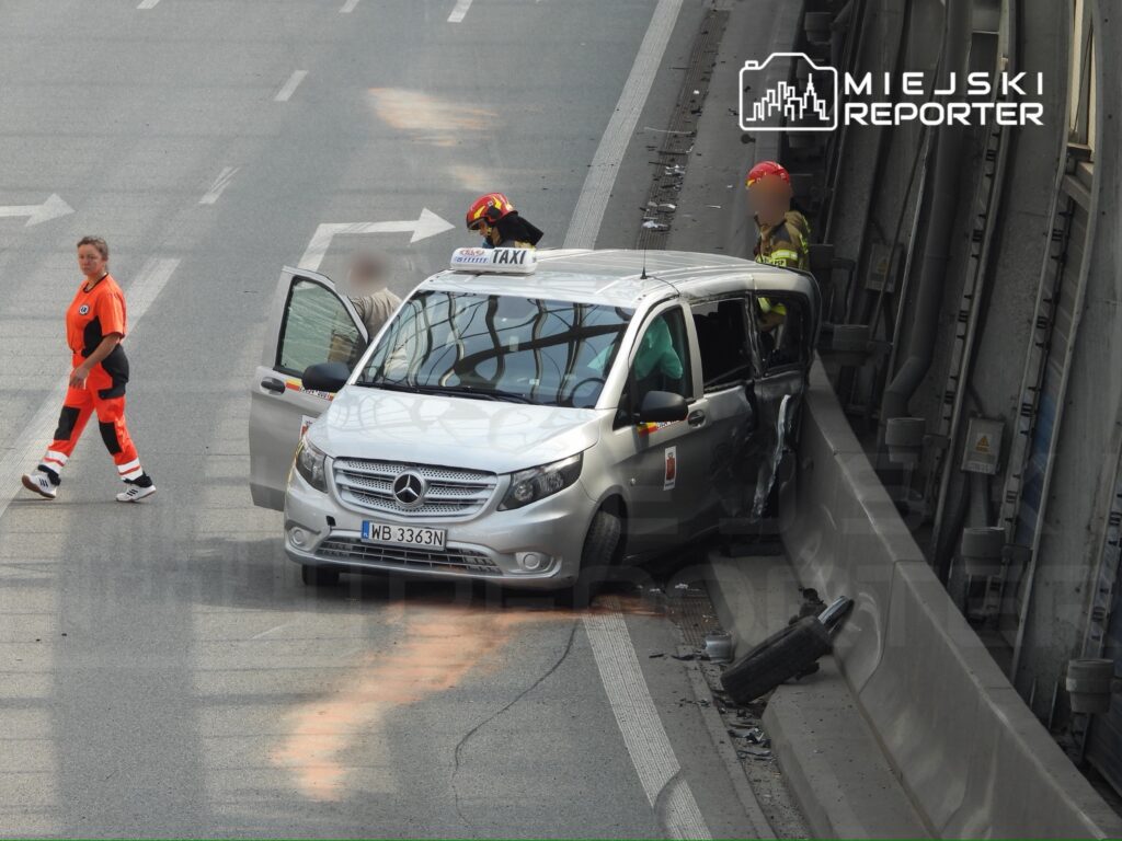 Taksi z uszkodzeniami stoi przy barierce na drodze, a ratownicy udzielają pomocy kierowcy.