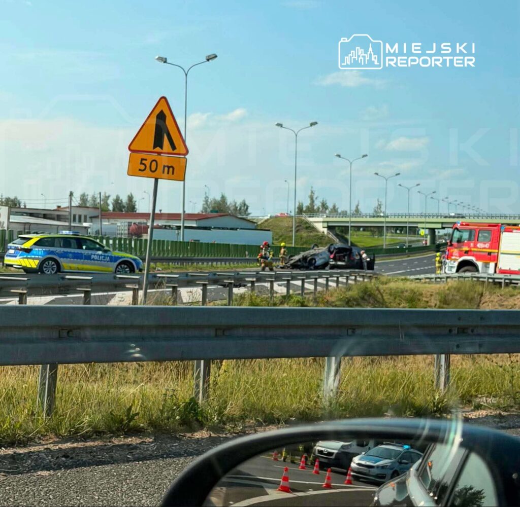 Na drodze leży przewrócony samochód, a obok interweniują strażacy i policja. W tle widoczny znak ostrzegawczy.