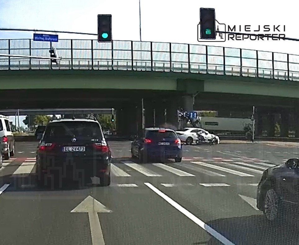 Ruchliwa ulica z sygnalizacją świetlną; czarne SUV-y i niebieski samochód oczekują na zielone światło.