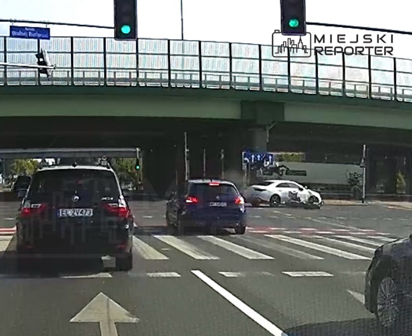 Ruch samochodów na skrzyżowaniu z sygnalizacją świetlną, w tle widoczny most i znaki drogowe.
