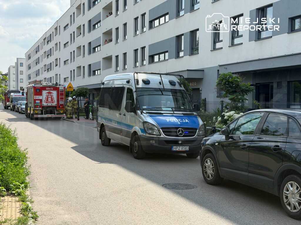 Wzdłuż ulicy parkują pojazdy policyjne i strażackie, a strażacy rozmawiają z funkcjonariuszami.