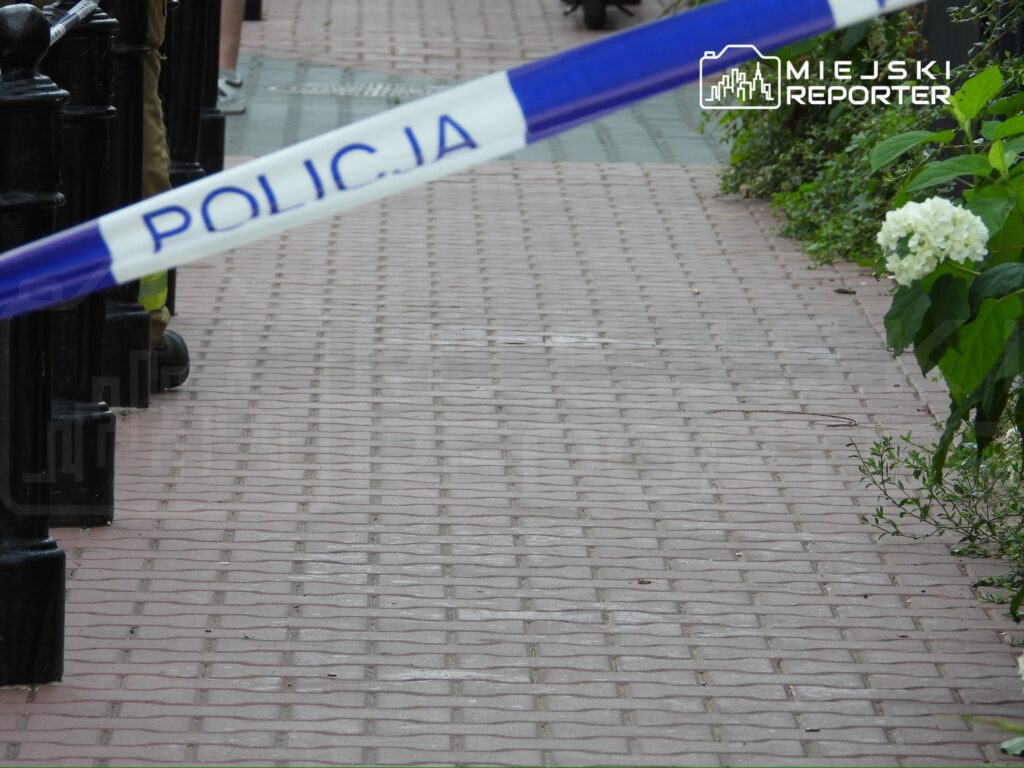 Na chodniku znajduje się policyjna taśma zabezpieczająca, a w tle widać osoby w mundurach oraz roślinność.