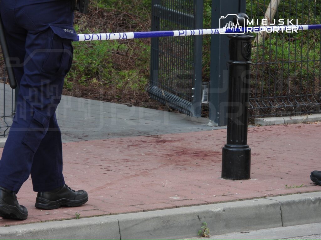 Policjant w mundurze stoi obok oznakowanego miejsca zdarzenia z plamą krwi na chodniku.