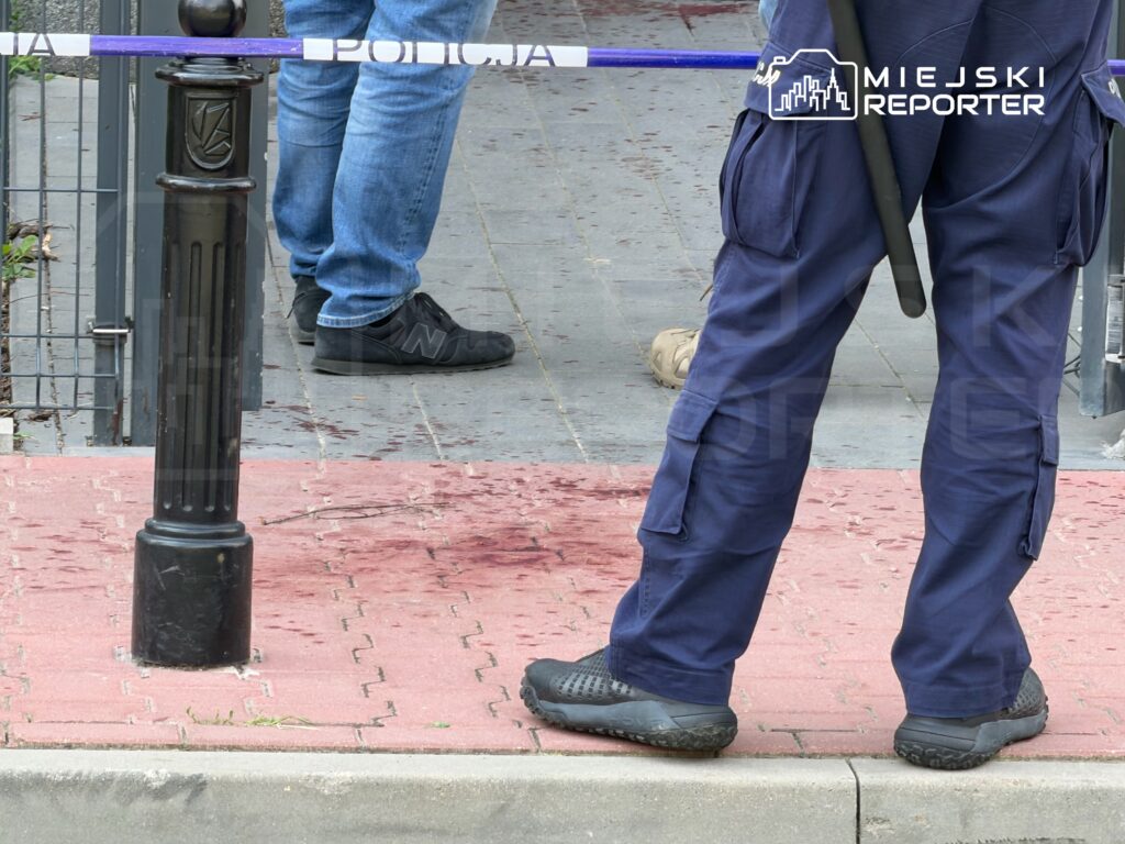 Funkcjonariusz policji stoi na chodniku, obok oznakowania miejsca zdarzenia, z widocznymi plamami na nawierzchni.
