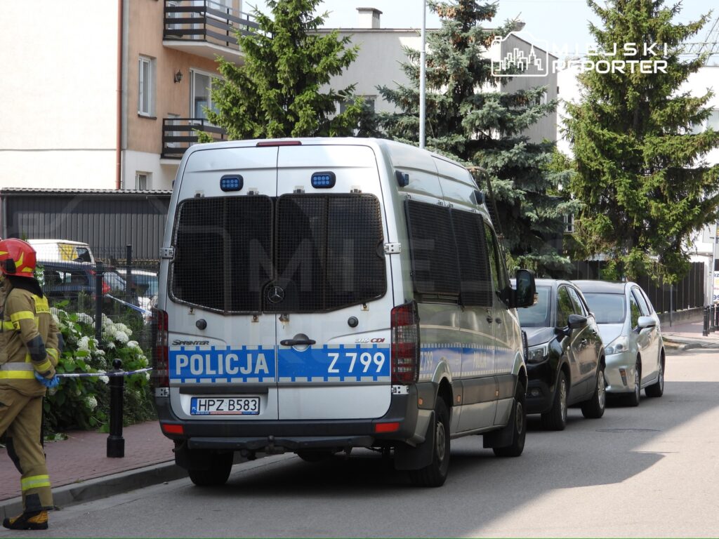 Policyjny radiowóz zaparkowany na ulicy, obok strażaka w hełmie, przy ogrodzeniu z kwiatami.