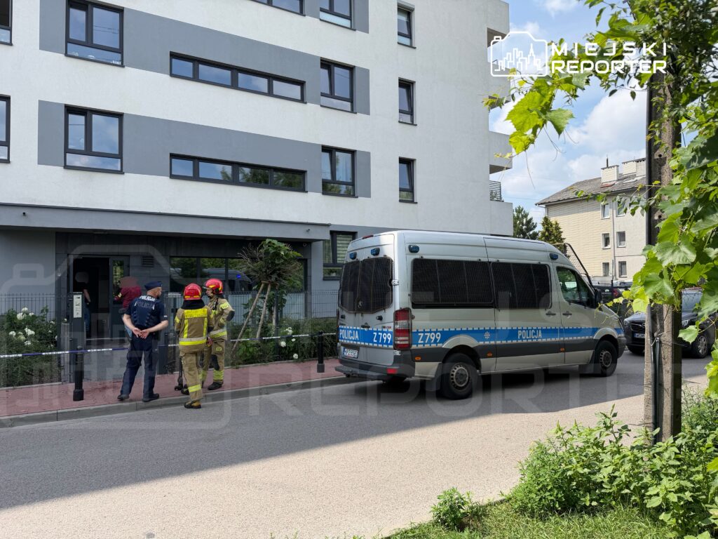 Policjant rozmawia z ratownikami w mundurach przed nowoczesnym budynkiem mieszkalnym, obok oznakowany radiowóz.