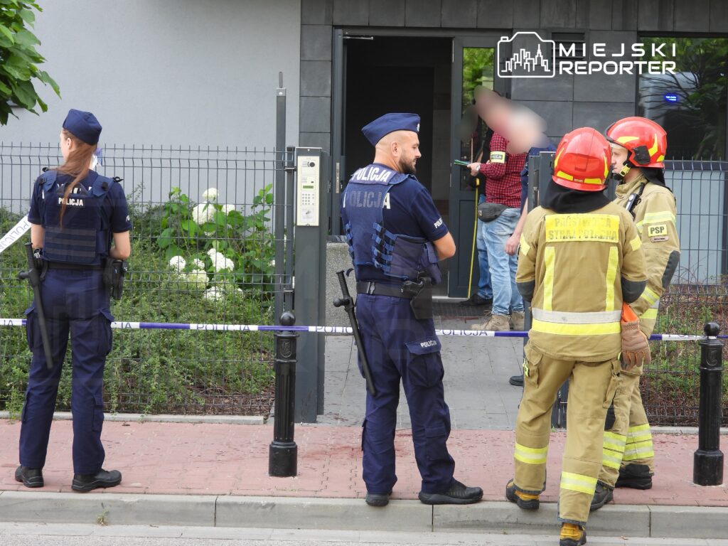 Grupa policjantów i strażaków rozmawia przed budynkiem, w tle widoczny mężczyzna z narzędziem w ręku.