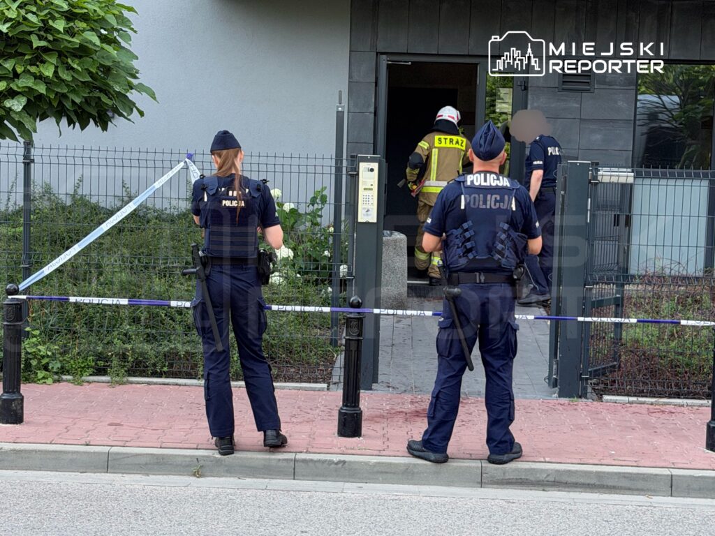 Policjanci zabezpieczają teren przed budynkiem, podczas gdy strażak wchodzi do środka.