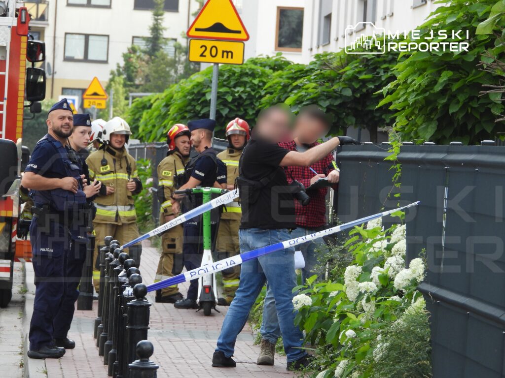 Grupa strażaków i policjantów stoi w pobliżu ogrodzenia, a jeden mężczyzna rozmawia z funkcjonariuszem.