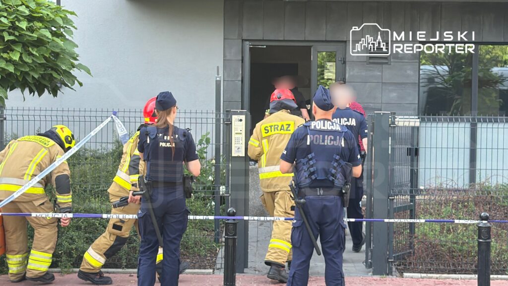 Grupa strażaków i policjantów wchodzi do budynku, zabezpieczonego taśmą policyjną.