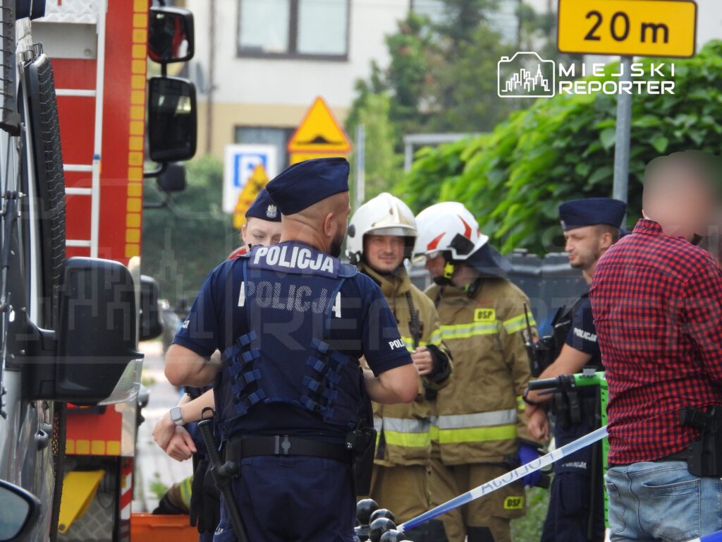 Grupa policjantów i strażaków rozmawia na ulicy, obok wozu strażackiego i oznakowania drogowego.